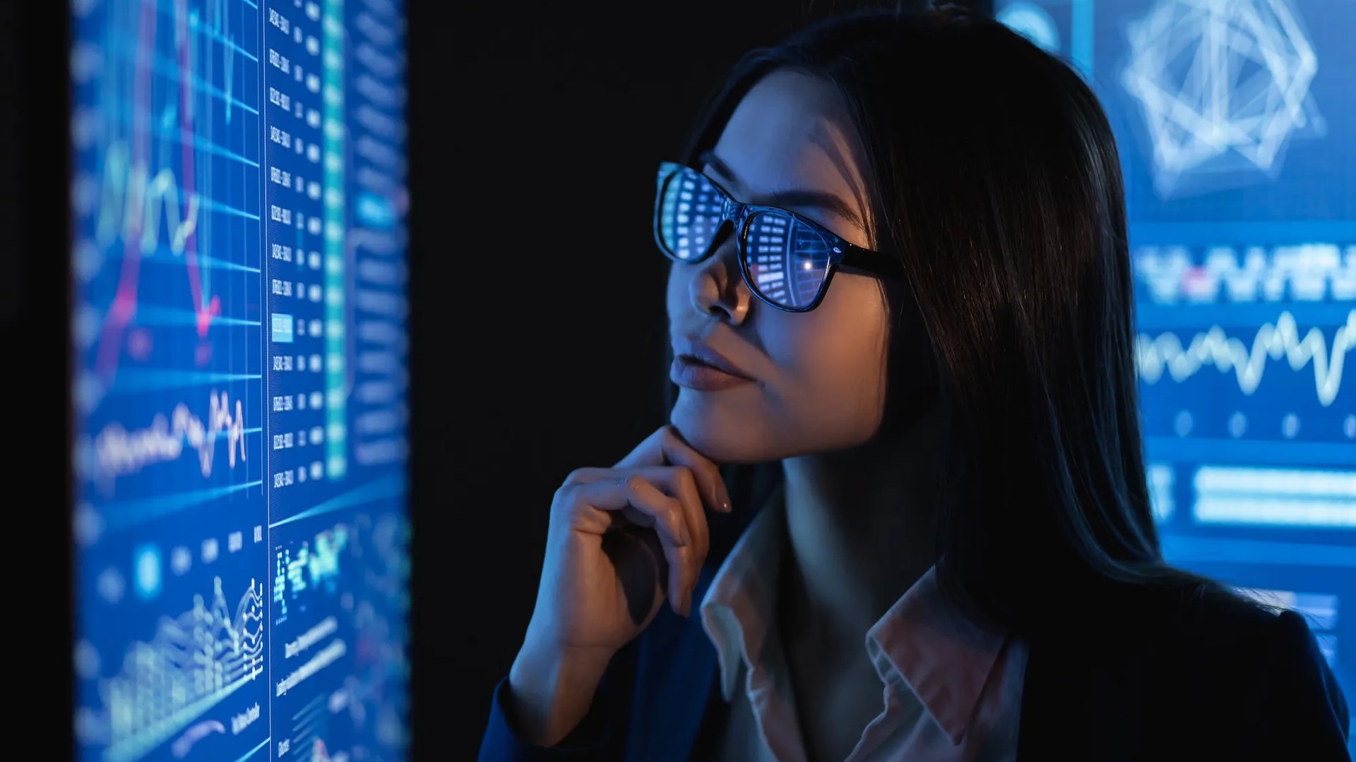 A woman wearing glasses looks thoughtfully at large illuminated data screens showing graphs, charts and complex visualisations. The blue light from the screens reflects in her glasses as she studies the information.