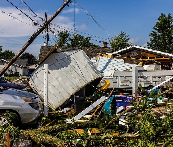 A residential street heavily damaged by a storm, with collapsed power lines, overturned vehicles and debris from destroyed buildings scattered across the ground.