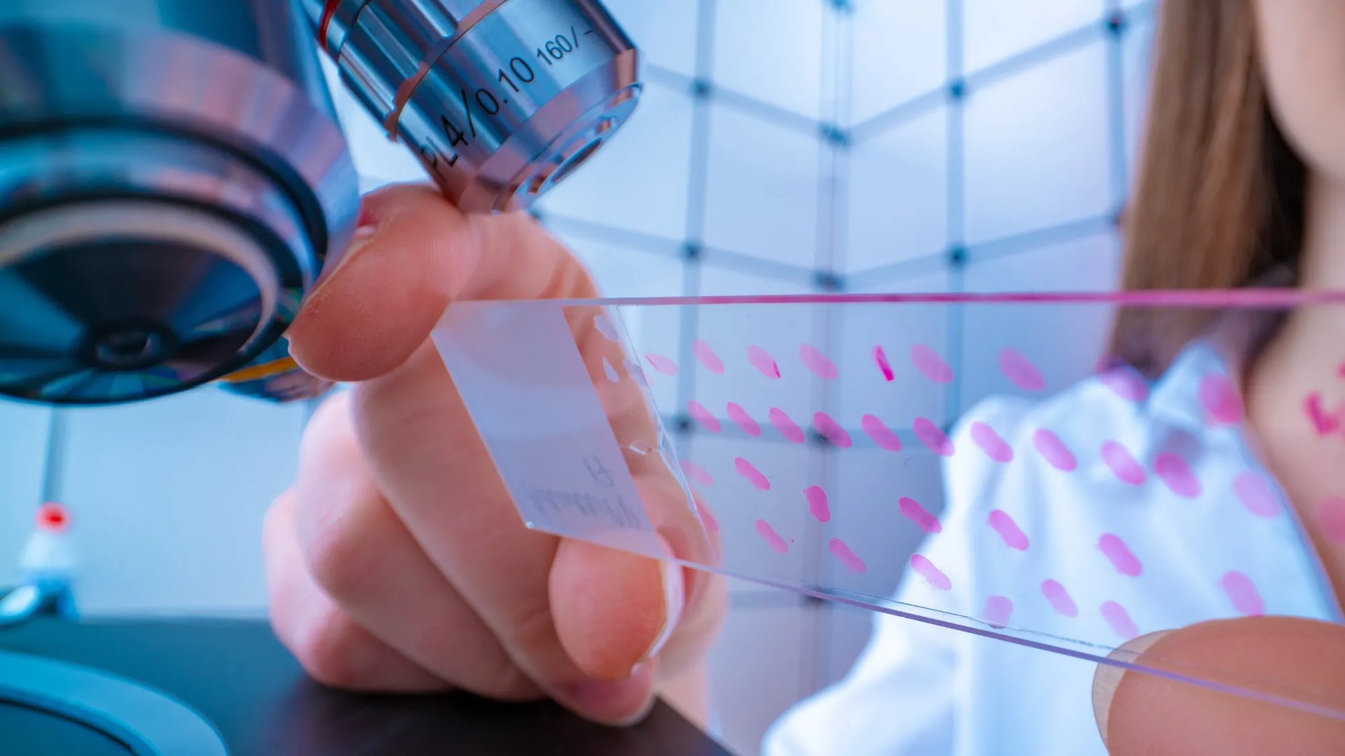 A researcher holds a glass slide marked with pink test samples under a microscope, examining it closely in a laboratory setting.