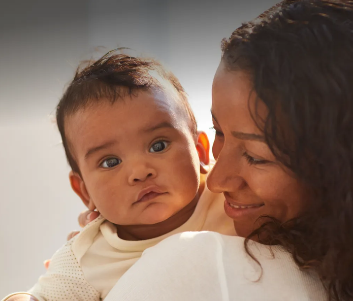 A mother smiles warmly while holding her baby, who looks directly at the camera with a calm expression.