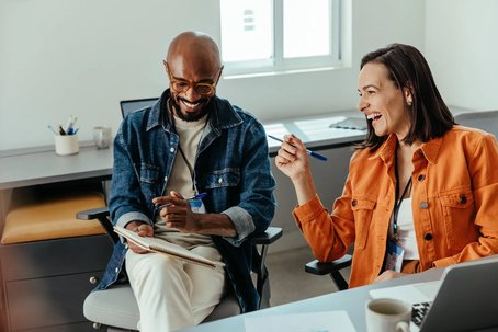 Two colleagues sit together in an office, smiling and discussing notes. One holds a notebook, the other a pen, and both appear engaged and relaxed in conversation.