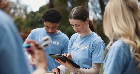 Charity volunteers in blue t-shirts working together outdoors, one using a tablet while others look on.