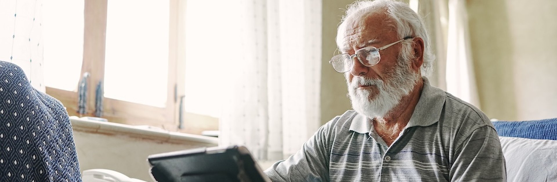 An elderly man with white hair and a beard sits on a patterned armchair, using a tablet device with a stand. He wears glasses and a striped polo shirt, concentrating on the screen.