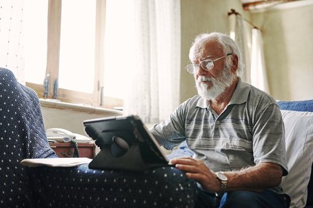 An elderly man with white hair and a beard sits on a patterned armchair, using a tablet device with a stand. He wears glasses and a striped polo shirt, concentrating on the screen.