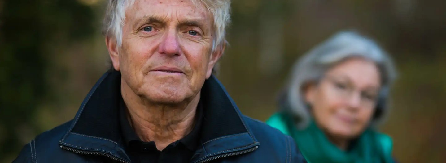 Older man in a black leather jacket standing outdoors with a serious expression, while an older woman in a green scarf looks on from the background, slightly out of focus.