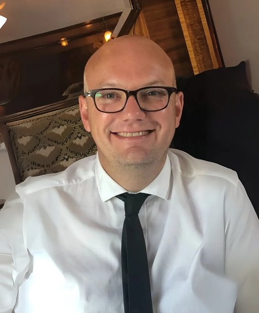 Peter is smiling on the Pullman train while celebrating his wedding anniversary, wearing glasses, a white shirt, and a black tie.