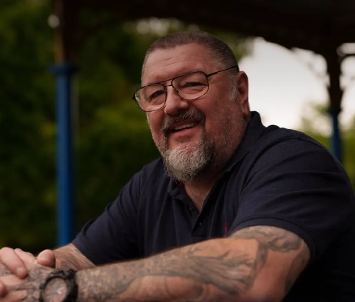 Smiling older man with glasses and tattoos on both arms, sitting outdoors under a shaded structure with greenery in the background.