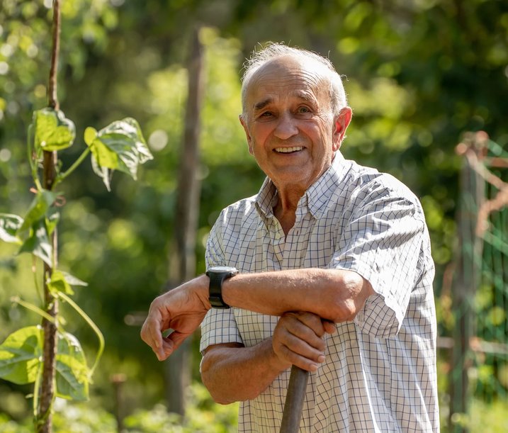 An elderly man smiling and leaning on a gardening tool in a lush, green garden on a sunny day.