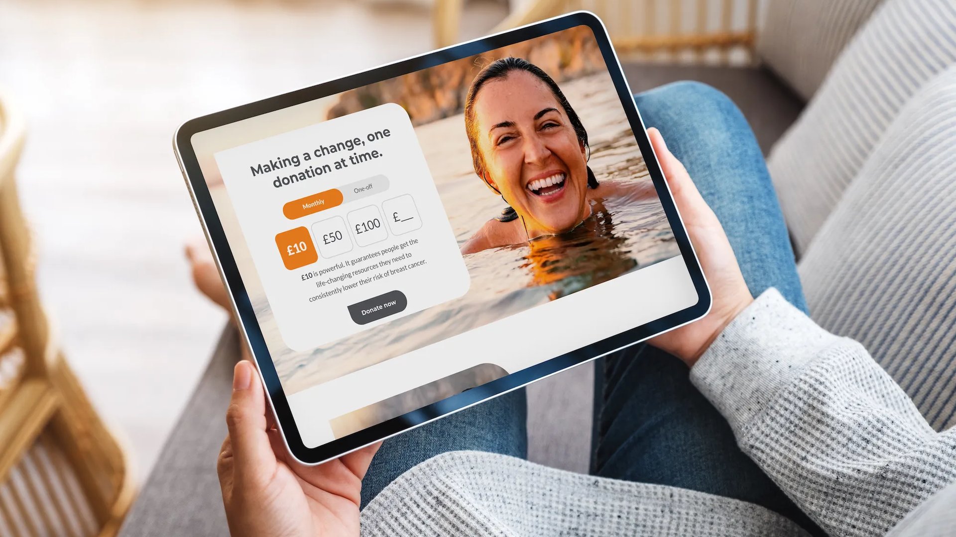 A person sitting on a sofa holding a tablet that displays a Breast Cancer UK donation page, featuring a smiling woman in the water and options to choose a monthly donation amount.