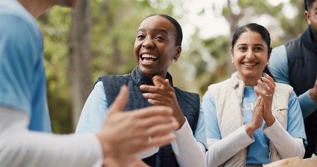 A group of volunteers outdoors smiling and clapping, with one woman in the centre laughing warmly while others around her join in.
