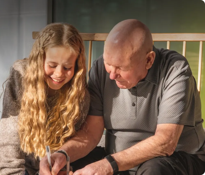 Smiling young girl and elderly man sitting together on a bench, writing or drawing in a notebook with natural sunlight streaming in.