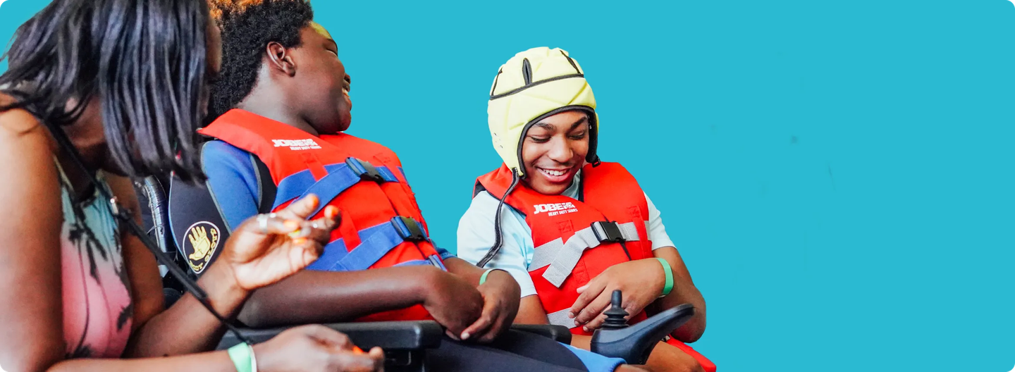Two children in wheelchairs wearing red life jackets and smiling, one wearing a yellow protective helmet, interacting joyfully with an adult.