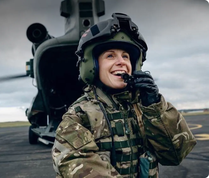 Smiling female soldier in camouflage uniform and helmet speaking into a radio, standing in front of a military helicopter on a tarmac.