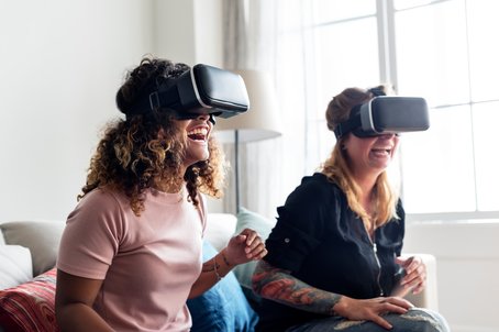 Two women laughing and enjoying a virtual reality experience while wearing VR headsets in a bright living room.