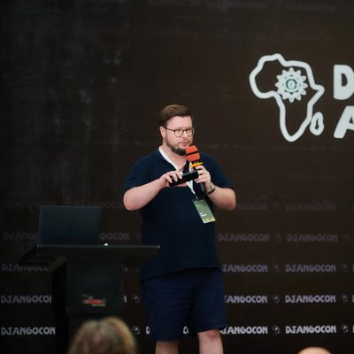 Jon speaking on stage at DjangoCon Africa, holding a microphone in front of a backdrop featuring the event’s logo and an outline of the African continent.