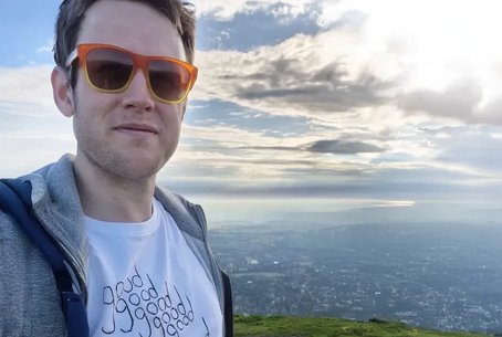 Jake stands on the Malvern Hills wearing orange sunglasses and a white T-shirt that reads “good good good good double good”, with a scenic view stretching out behind him under a dramatic cloudy sky.