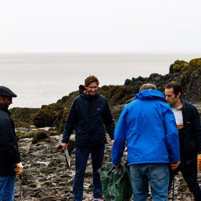 Giant Digital team members taking part in a beach clean, standing on a rocky shoreline wearing gloves and outdoor clothing while collecting litter.