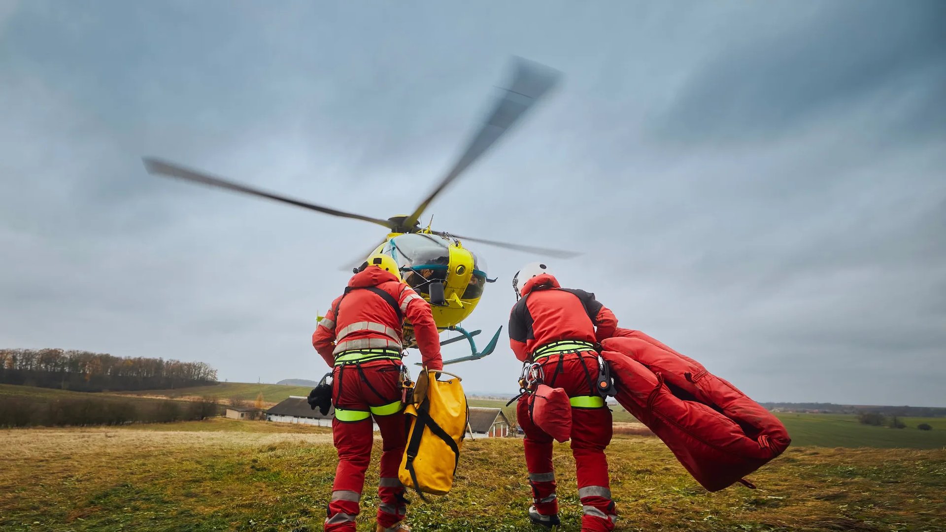Two emergency responders in red flight suits carry equipment towards a yellow rescue helicopter preparing for take-off on a cloudy day, symbolising teamwork and urgent response in life-saving missions.