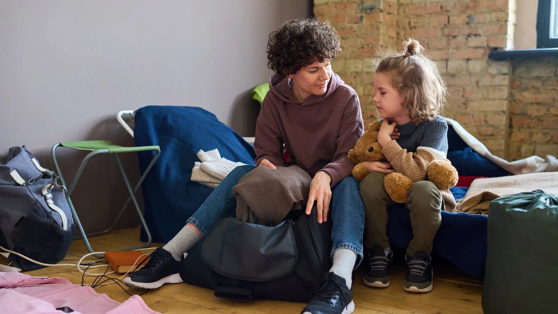 A parent and child sit together on makeshift bedding in a shelter, the parent packing clothes into a bag while the child holds a teddy bear.