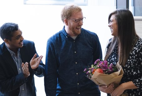 Three Giant Digital colleagues smiling and celebrating together in a bright office space. One person holds a bouquet of flowers, while another claps in appreciation.