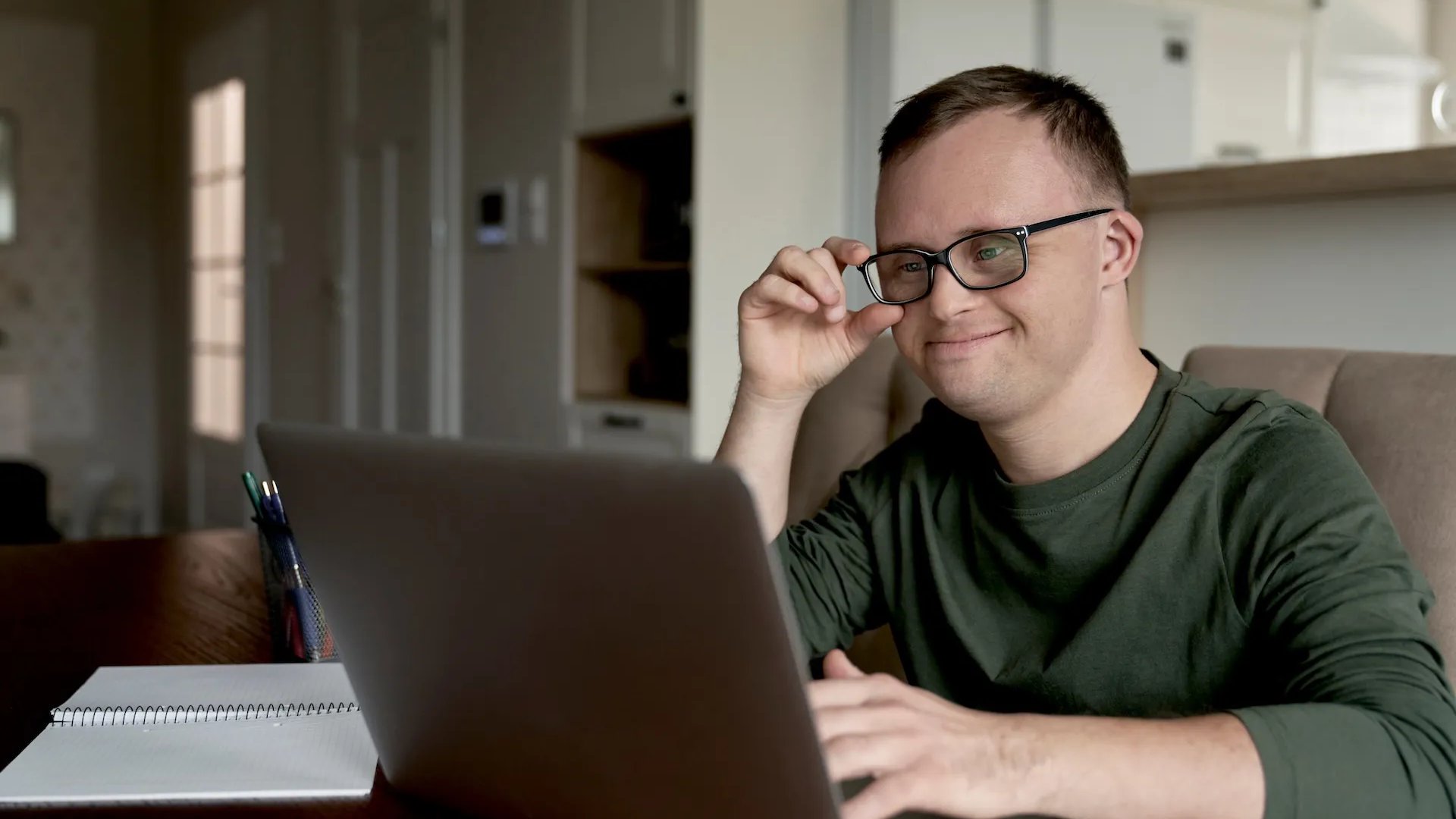 A young man with Down syndrome wearing glasses sits at a table using a laptop, smiling as he adjusts his glasses. A notebook and pens are on the table beside him.