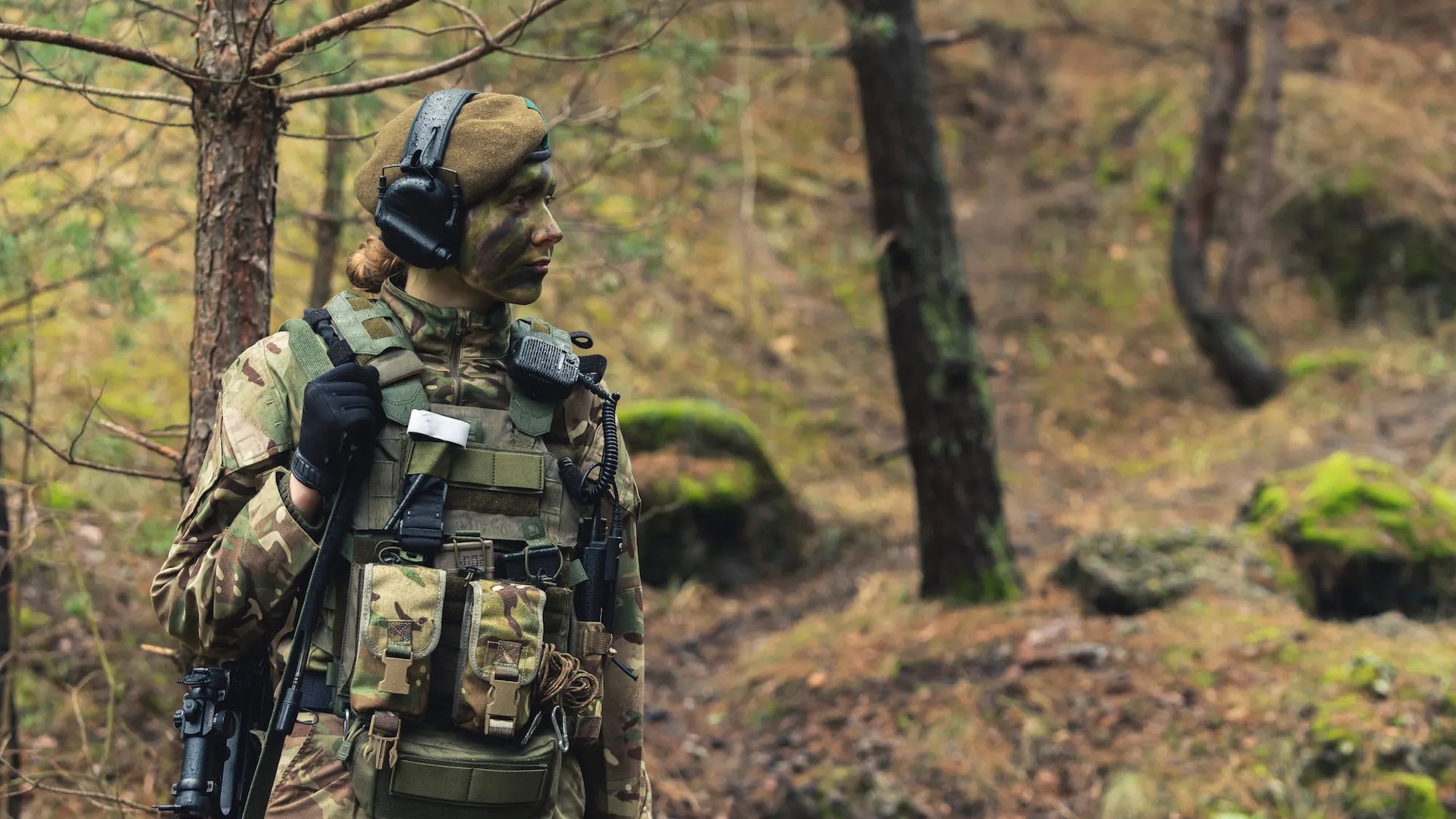 Member of the British Armed Forces standing in woodland, wearing camouflage uniform and radio headset.