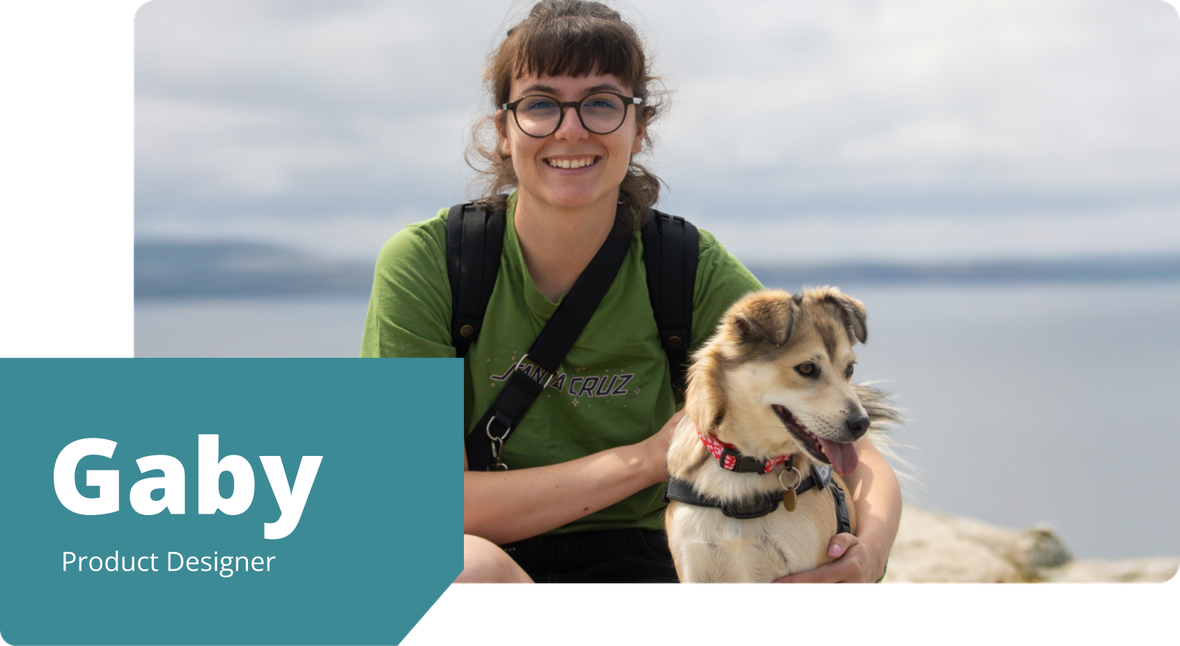 Gaby, wearing glasses and a green t-shirt, smiles while sitting outdoors with her rescue dog Maya, overlooking a scenic body of water.
