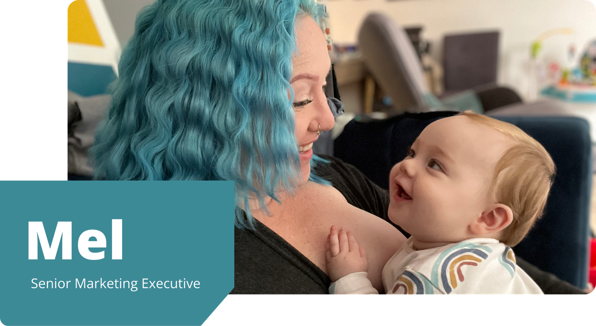 Mel, with bright blue hair, smiles warmly at her baby son in a rainbow-patterned outfit, as they share a joyful moment at home.
