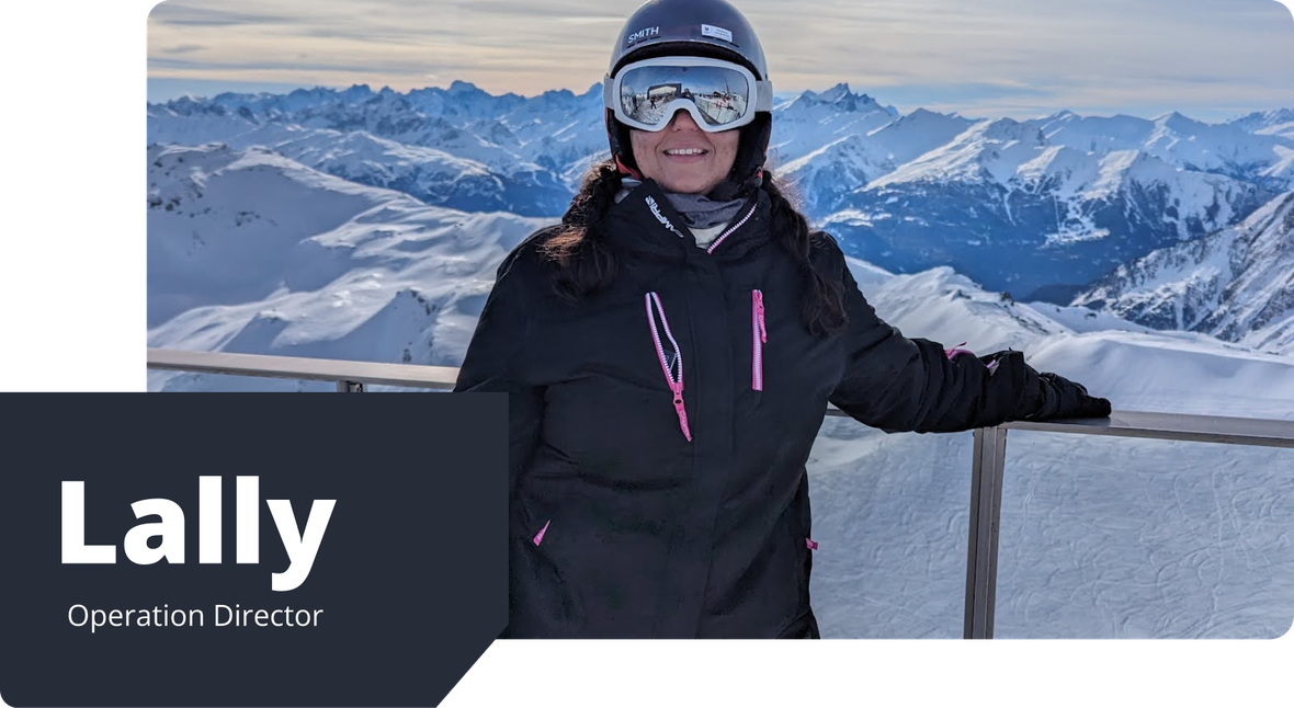 Lally, wearing a black ski jacket, helmet and goggles, smiles while standing against a snowy mountain backdrop at a ski resort.