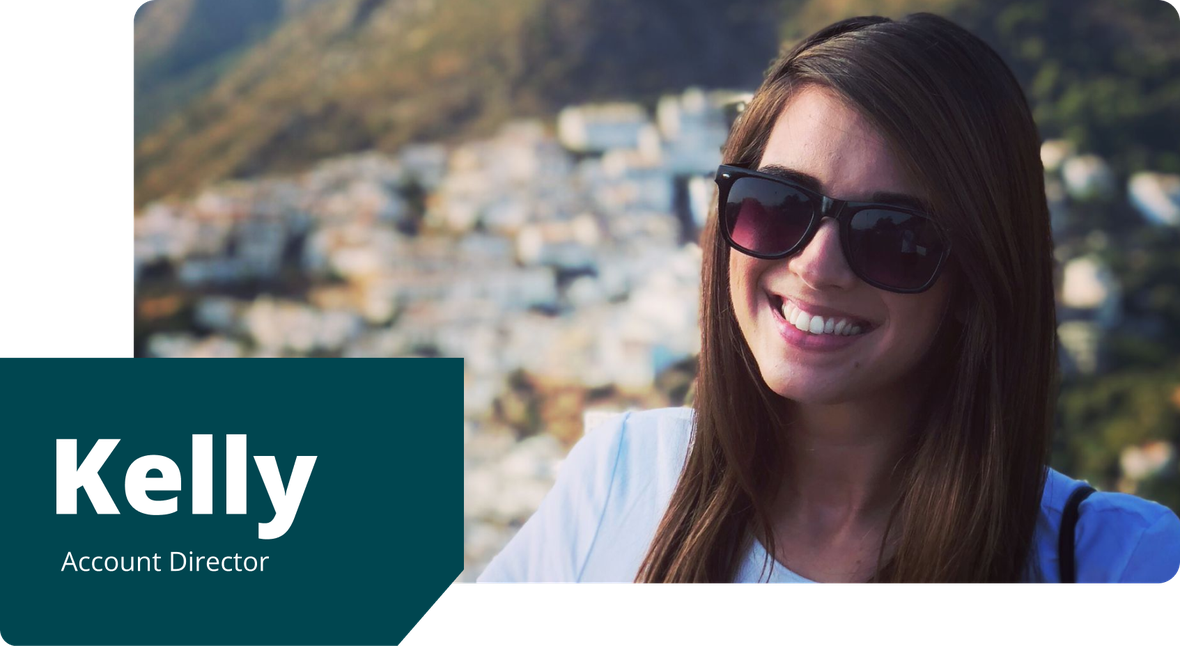 Kelly is smiling and wearing sunglasses, standing outdoors with a scenic Spanish hillside town and mountains in the background.
