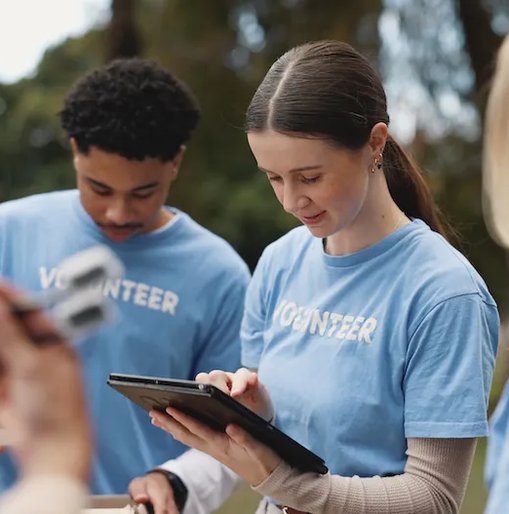 Charity volunteers in blue t-shirts working together outdoors, one using a tablet while others look on.