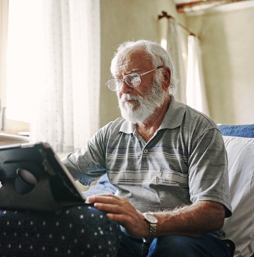 An elderly man with white hair and a beard sits on a patterned armchair, using a tablet device with a stand. He wears glasses and a striped polo shirt, concentrating on the screen.