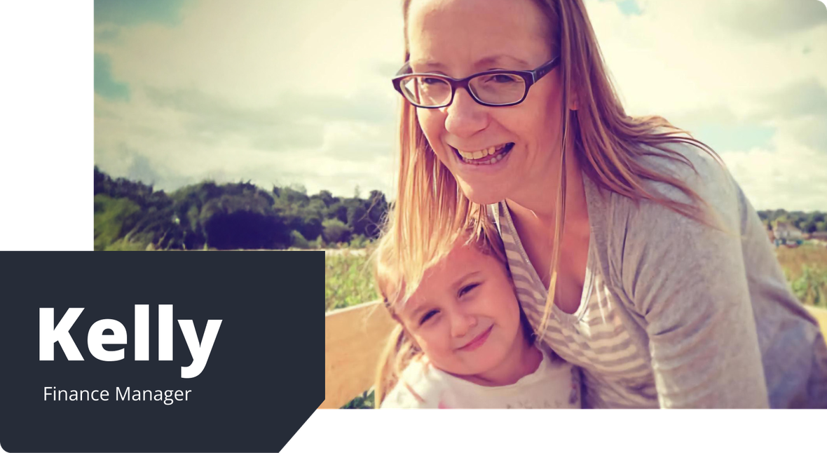 Kelly, wearing glasses and striped top, smiles while cuddling her daughter in a field, both enjoying a sunny day outdoors.
